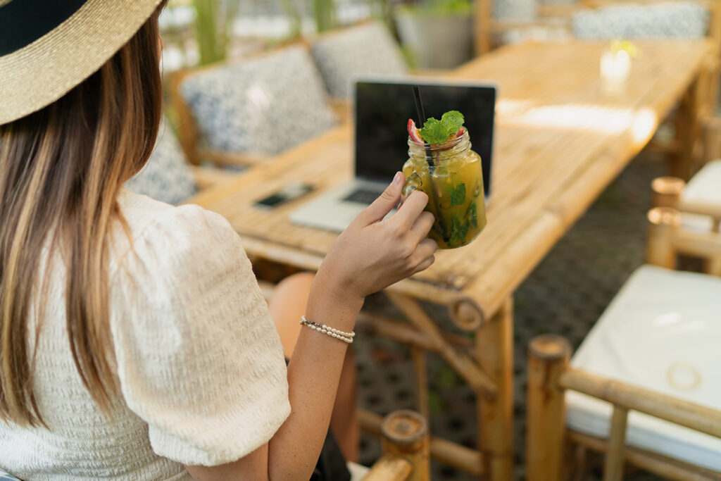 Pretty woman in straw hat using lap top while traveling in asia. Working remotely in stylish tropical cafe. Holding exotic beverage.