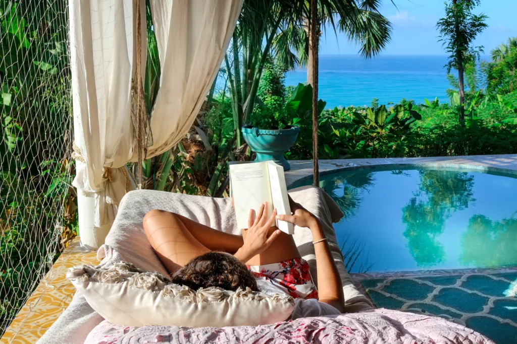 A beautiful shot of a person lying on chaise lounge reading a book near a pool with tropical plants - bali dive resort