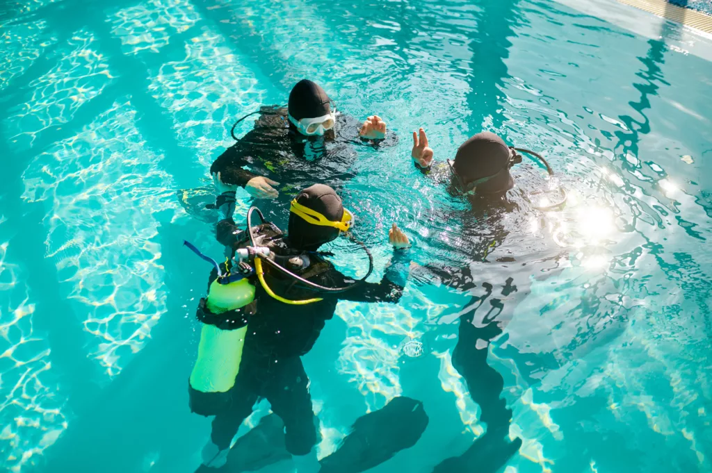 Divemaster and two divers in aqualungs, course in diving school. Teaching people to swim underwater with scuba gear, indoor swimming pool interior on background, group training - Bali Dive Resort - Bali Scuba Diving