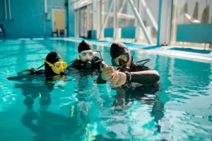 Instructor and two divers in aqualungs, dive lesson in diving school. Teaching people to swim underwater with scuba gear, indoor swimming pool interior on background, group training - Bali Diving Courses
